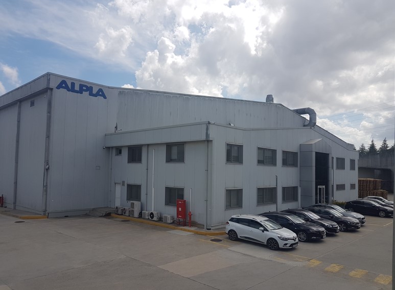 Exterior view of the ALPLA production facility in Yüreğir, Türkiye, showcasing an industrial building with parking spaces and the ALPLA logo prominently displayed on the factory wall.